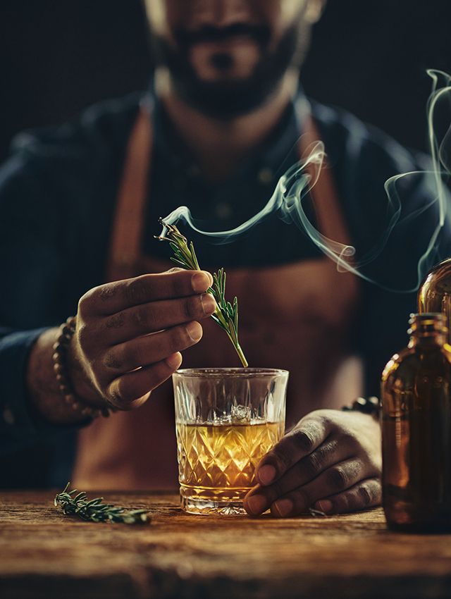 bartender putting a smoking piece of rosemary into a glass filled with amber liquid